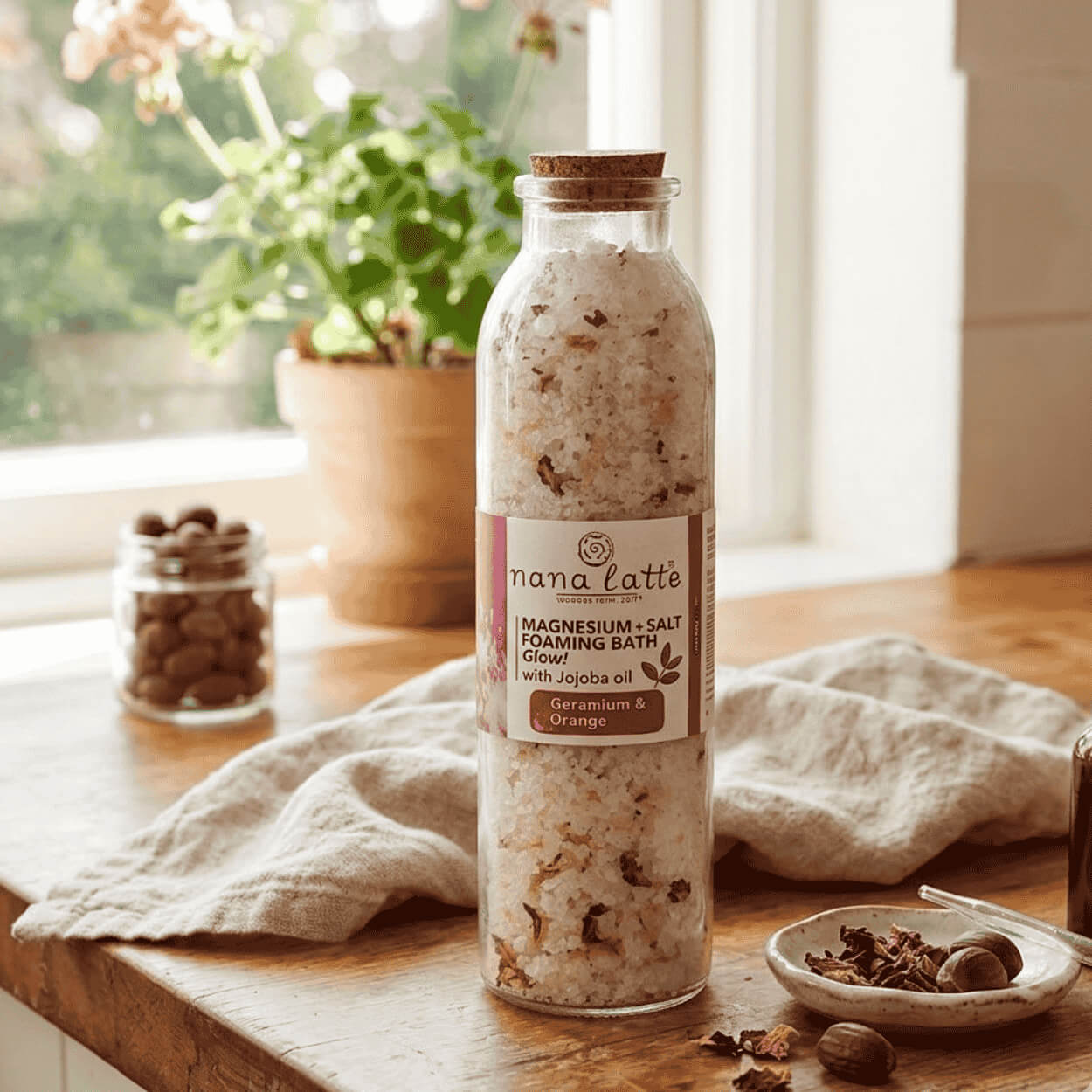 Produktová fotografie zobrazuje skleněnou lahev koupelové soli Magnesium + Salt Foaming Bath Glow! Geranium & Orange na dřevěné ploše u okna.